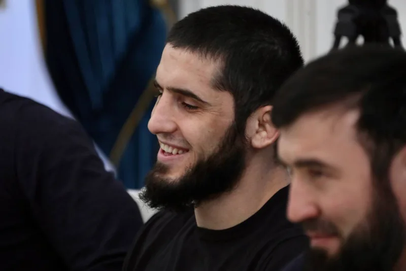 Islam Makhachev smiling in a black T-shirt, seated beside a bearded man indoors
