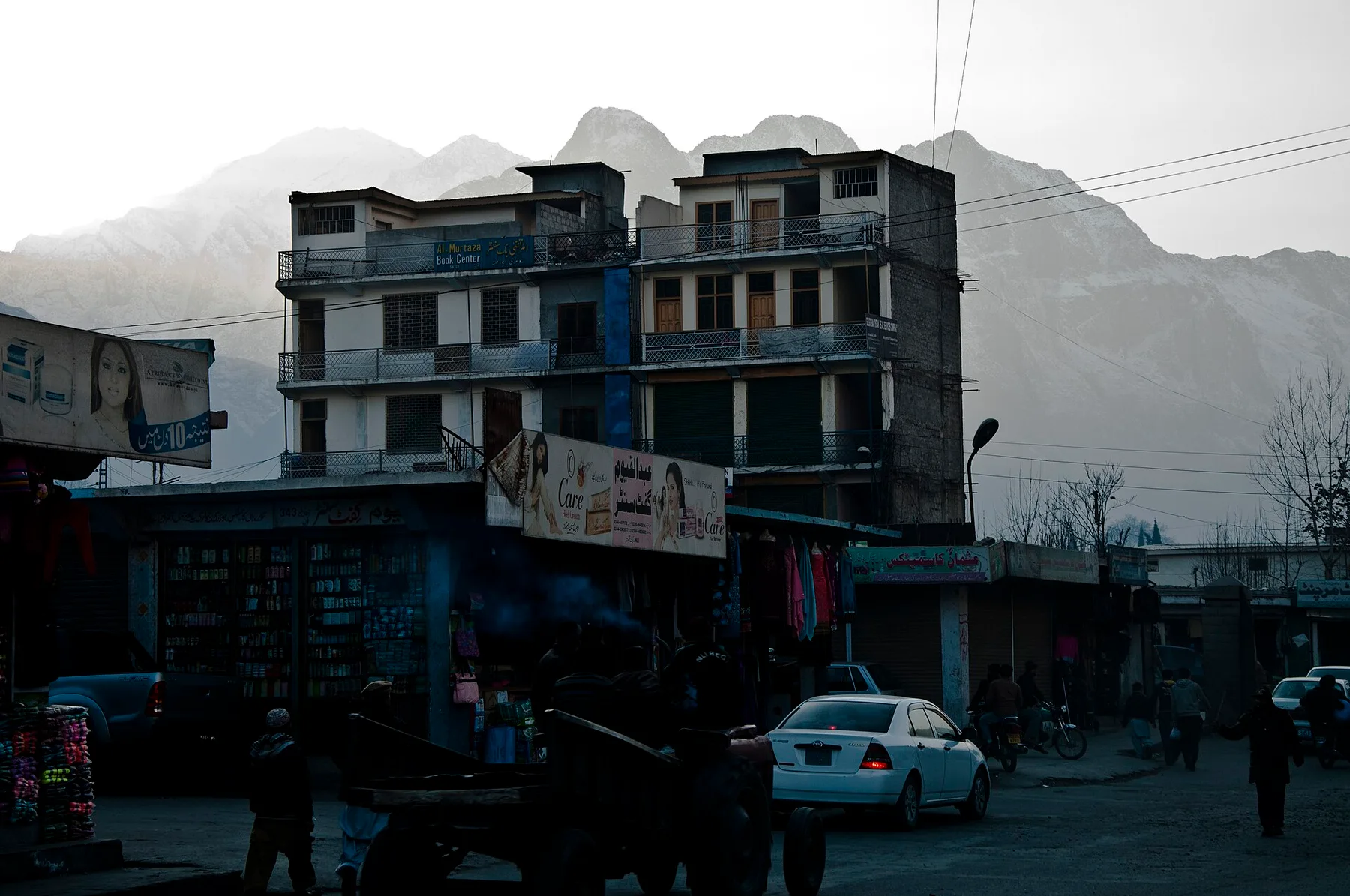 Street in Gilgit with shops, a white car and a multi-storey building below snow-capped mountains