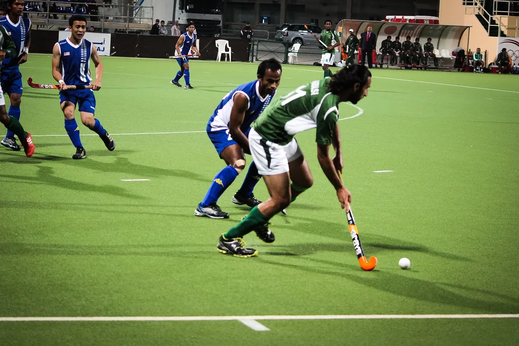 Pakistan hockey player in green dribbling the ball past a defender in blue on an artificial pitch
