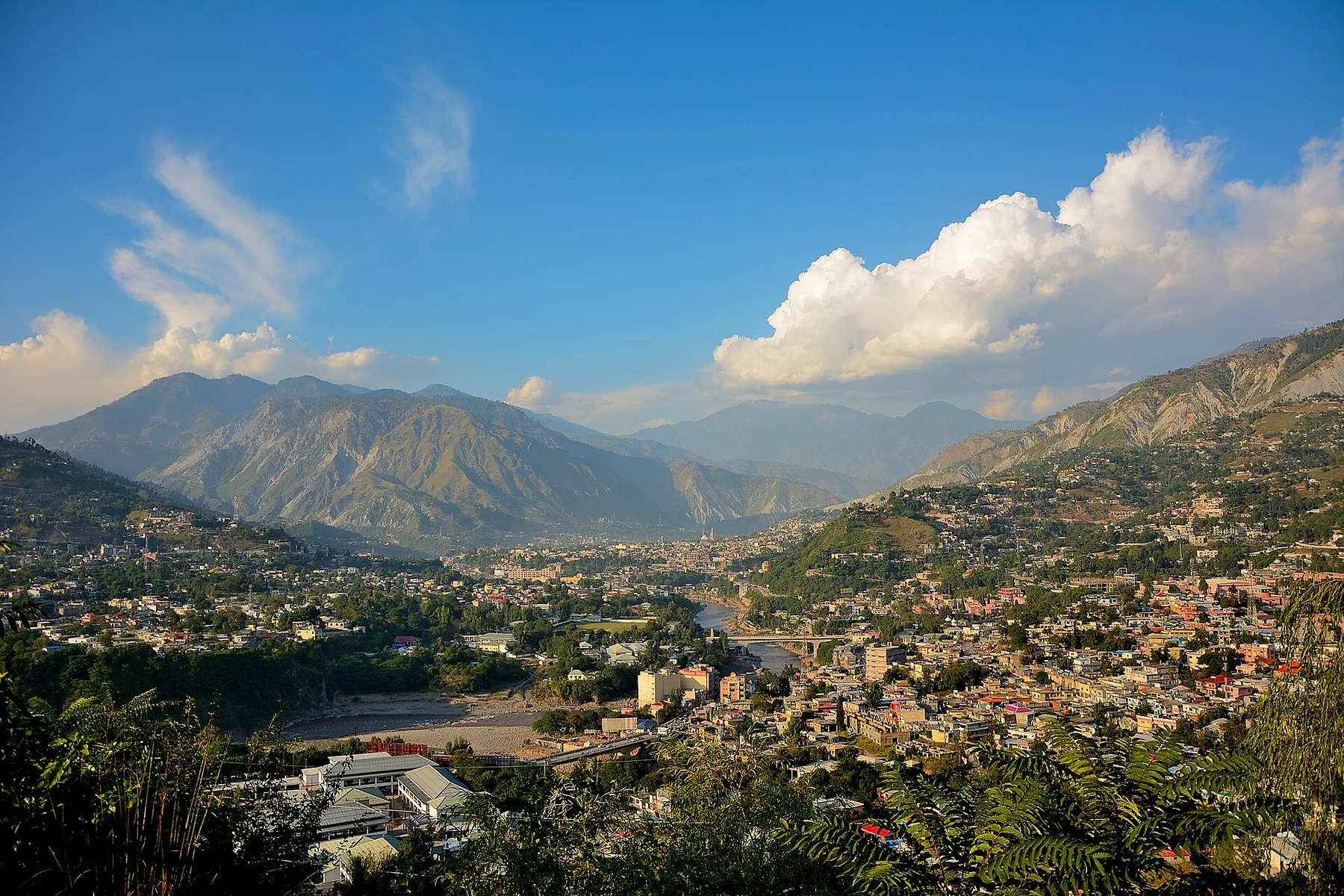 Muzaffarabad city spread along a river valley beneath steep mountains under a blue sky