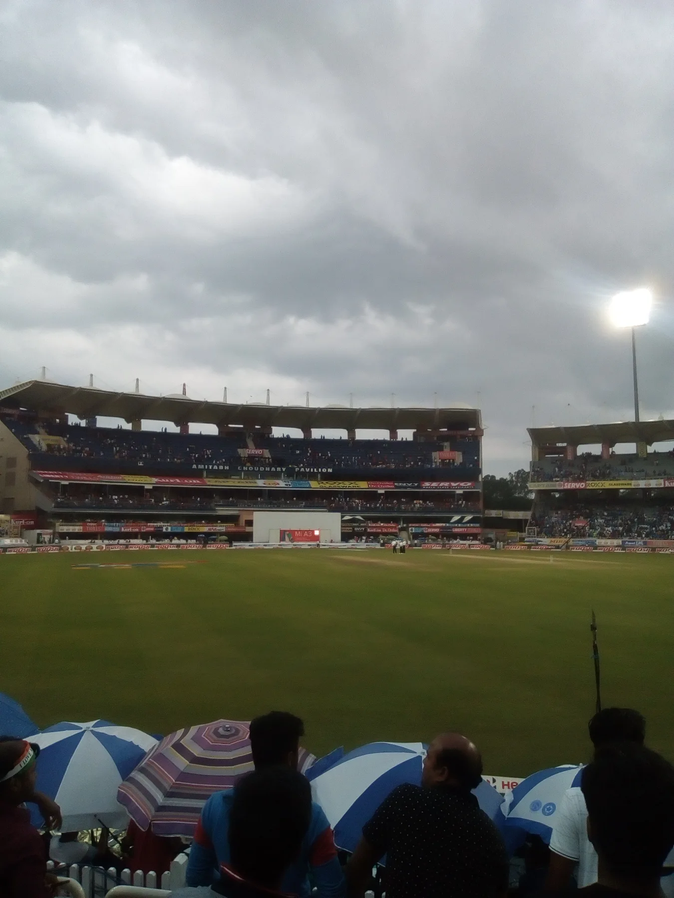 Cricket ground under grey clouds with floodlights on and fans holding umbrellas in the stand
