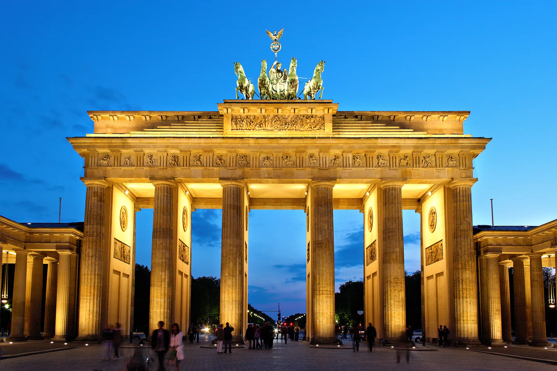 The Brandenburg Gate in Berlin lit up at dusk, with people walking in the square in front of it
