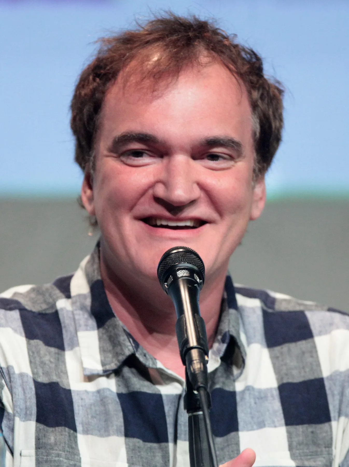 Quentin Tarantino smiling at a microphone in a blue and white checked shirt