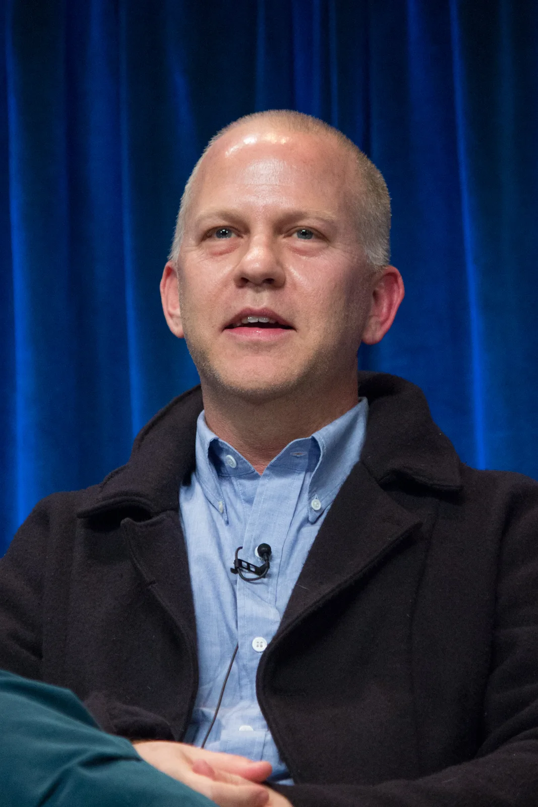 Ryan Murphy in a dark coat and blue shirt speaking on a panel in front of a blue curtain