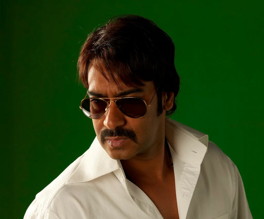 Ajay Devgn in aviator sunglasses and a white open-collared shirt against a green backdrop