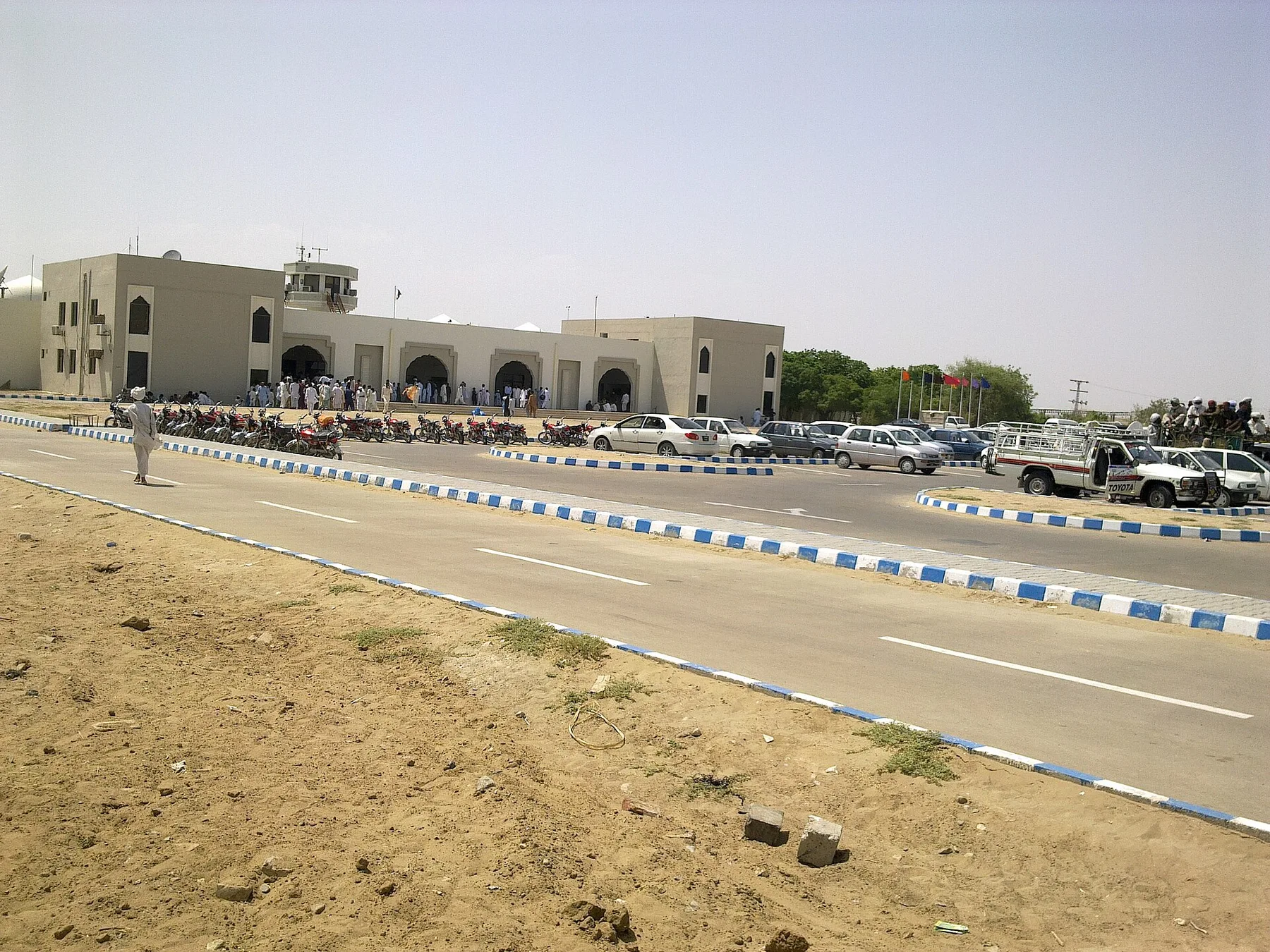 Single-storey airport terminal with a control tower, cars parked and travellers waiting outside on a dry sunny day