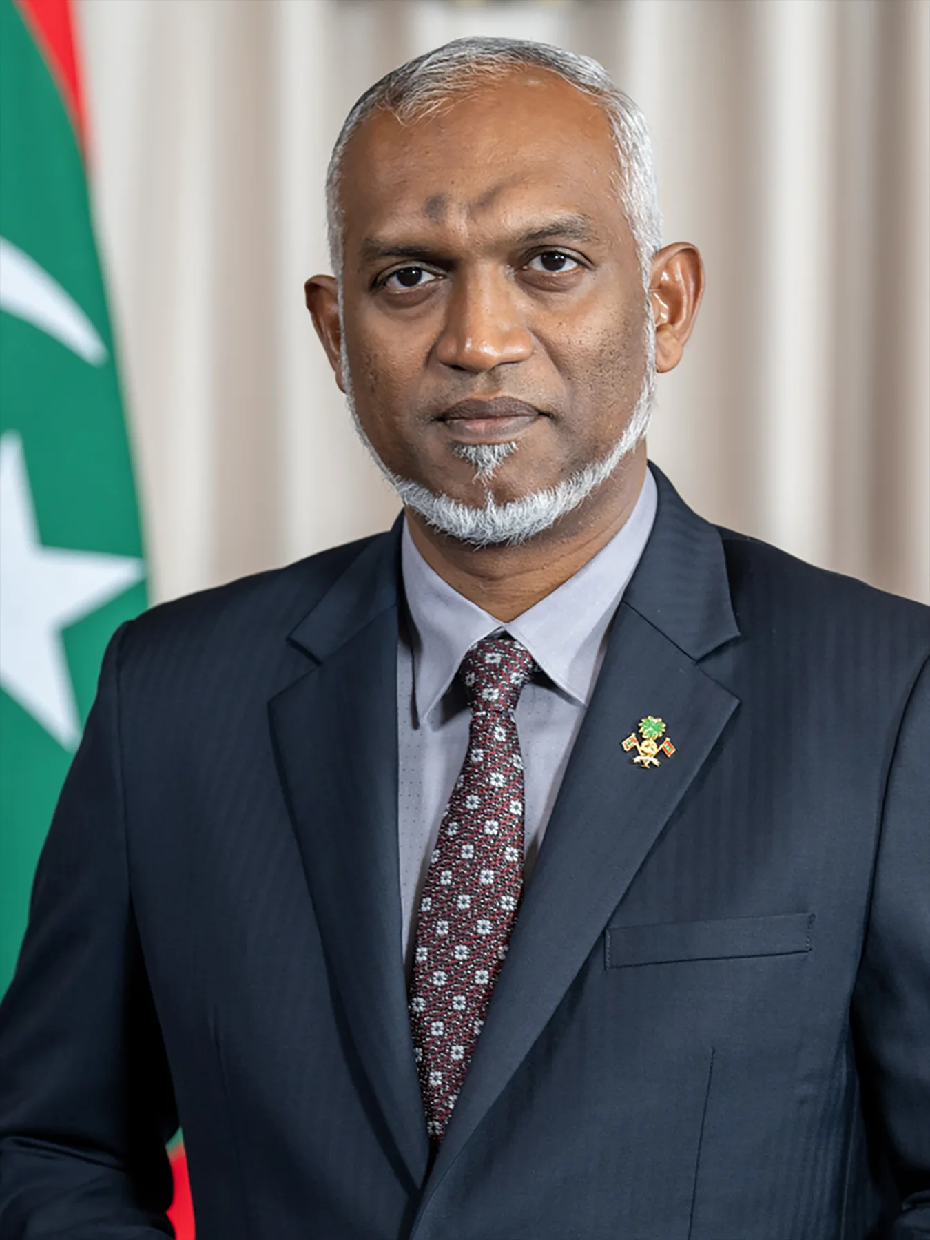 Maldives President Mohamed Muizzu in a dark suit and patterned tie, standing before the Maldivian flag