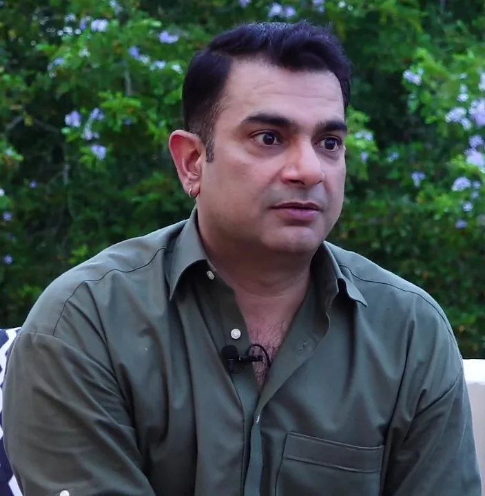 Sarmad Khoosat in an olive green shirt with a clip-on microphone, seated outdoors before flowering shrubs